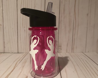 May include: Pink water bottle with a black lid and a white silhouette of two ballerinas on the side. The bottle has a straw.
