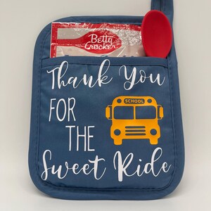 Puede incluir: Manopla de horno azul con el texto "Thank You For The Sweet Ride" y una ilustraci&oacute;n de autob&uacute;s escolar amarillo. Una cuchara roja y una bolsa transparente con el logotipo de Betty Crocker est&aacute;n en el bolsillo de la manopla.