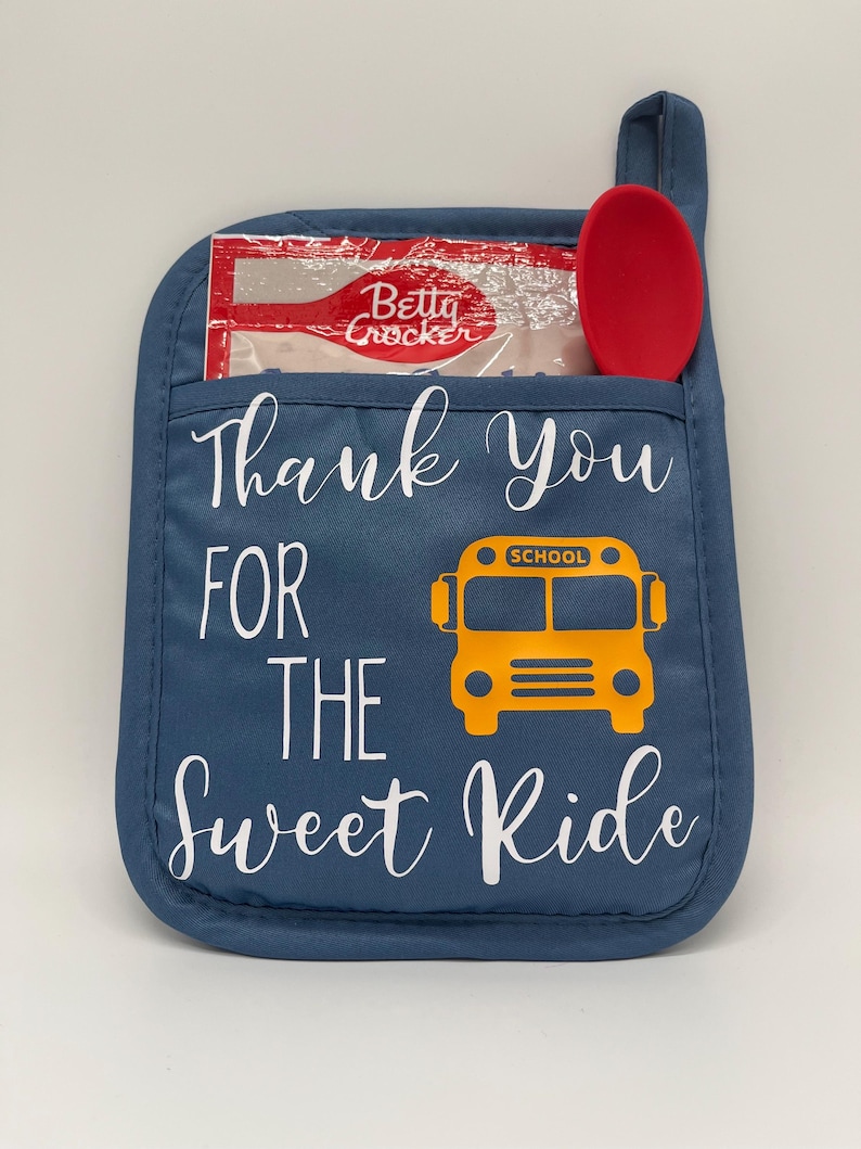 Puede incluir: Manopla de horno azul con el texto "Thank You For The Sweet Ride" y una ilustraci&oacute;n de autob&uacute;s escolar amarillo. Una cuchara roja y una bolsa transparente con el logotipo de Betty Crocker est&aacute;n en el bolsillo de la manopla.