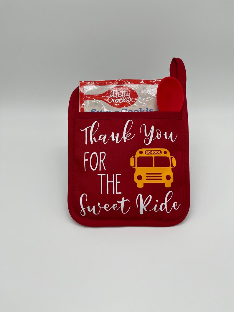 Puede incluir: Manopla de horno roja con el texto "Thank You For The Sweet Ride" y un gr&aacute;fico de autob&uacute;s escolar amarillo. Una peque&ntilde;a cuchara roja y un paquete de galletas de az&uacute;car Betty Crocker est&aacute;n dentro de la manopla.