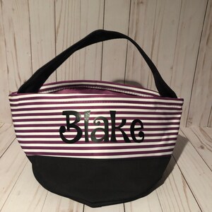 May include: Purple and white striped tote bag with a black handle and a black bottom. The bag has the name "Blake" written on the front in black.