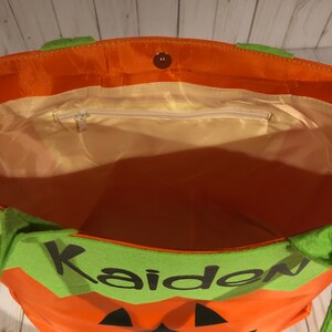 May include: Orange and green Halloween trick-or-treat bag with a pumpkin face and the name "Kaiden" on the front. The bag has a zippered pocket on the inside.