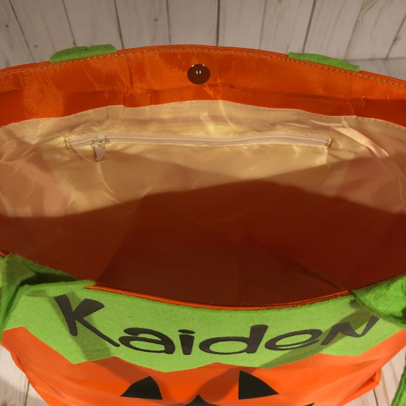 May include: Orange and green Halloween trick-or-treat bag with a pumpkin face and the name "Kaiden" on the front. The bag has a zippered pocket on the inside.