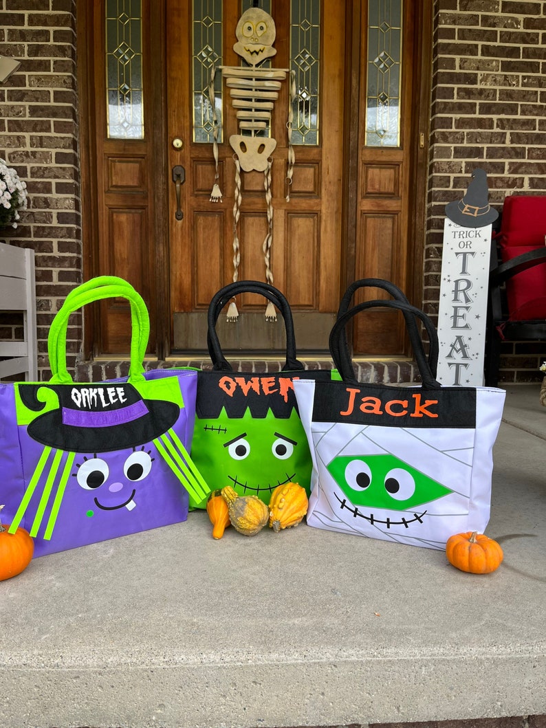 May include: Three Halloween trick-or-treat bags with personalized names: Oakley, Owen, and Jack. The bags are decorated with different Halloween characters: a witch, a Frankenstein monster, and a mummy. The bags are sitting on a concrete porch with a pumpkin in front of each bag.