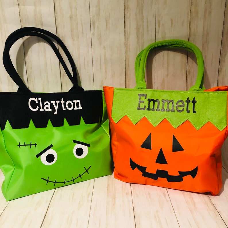 May include: Two Halloween trick-or-treat bags. One is black with a green Frankenstein face and the name Clayton. The other is orange with a pumpkin face and the name Emmett.