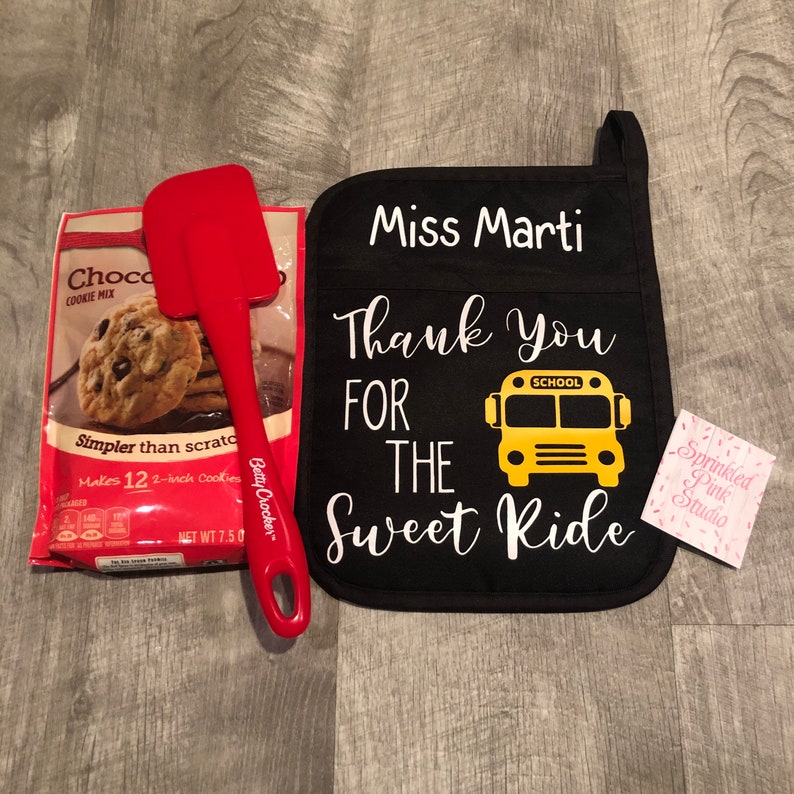 Puede incluir: Manopla de horno negra con el texto "Miss Marti Thank You For The Sweet Ride" y un gr&aacute;fico de autob&uacute;s escolar amarillo. La manopla de horno est&aacute; acompa&ntilde;ada de una esp&aacute;tula roja y una bolsa de mezcla para galletas con chispas de chocolate.