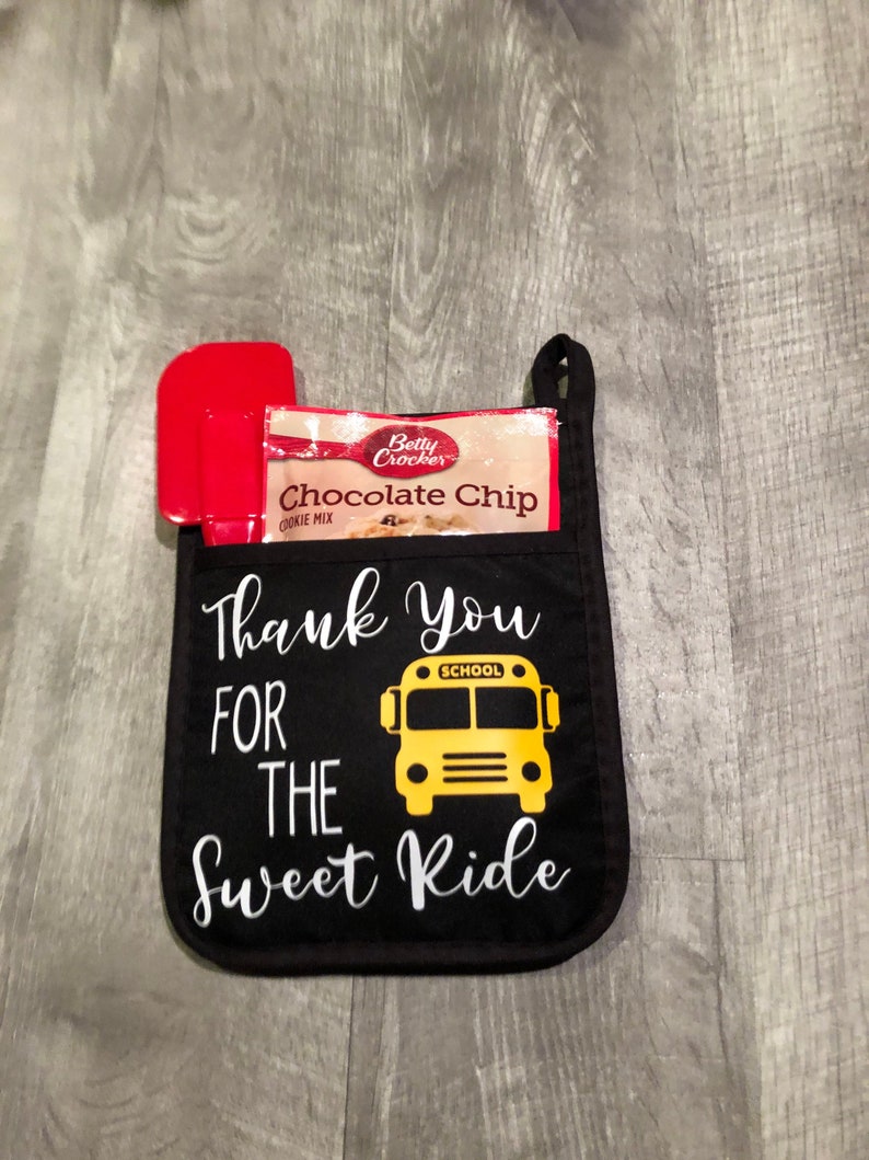 Puede incluir: Manopla de horno negra con el texto "Thank You For The Sweet Ride" y un gr&aacute;fico de autob&uacute;s escolar amarillo. La manopla de horno tiene una esp&aacute;tula roja y un paquete de mezcla para galletas con chispas de chocolate Betty Crocker.