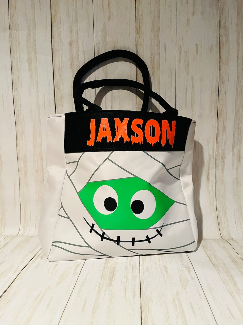 May include: White canvas tote bag with a mummy face design. The mummy has green eyes and a stitched mouth. The bag has a black fabric handle and a black fabric panel with the name "Jaxson" in orange dripping letters.
