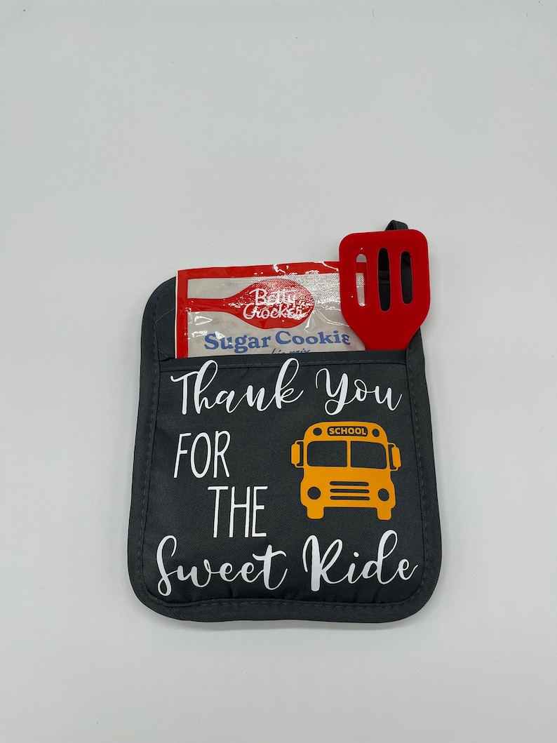 Puede incluir: Manopla de horno gris con el texto "Thank You For The Sweet Ride" y un gr&aacute;fico de autob&uacute;s escolar amarillo. Una esp&aacute;tula roja y un paquete de mezcla para galletas de az&uacute;car Betty Crocker est&aacute;n en el bolsillo de la manopla.