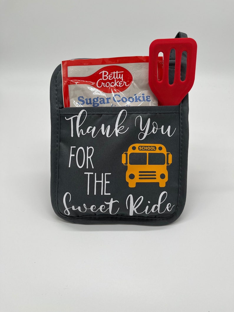 Puede incluir: Manopla de horno gris con el texto "Thank You For The Sweet Ride" y un gr&aacute;fico de autob&uacute;s escolar amarillo. Una esp&aacute;tula roja y un paquete de mezcla para galletas Betty Crocker Sugar Cookie est&aacute;n en el bolsillo de la manopla de horno.