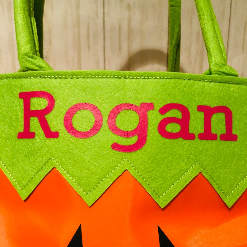 May include: Green felt trick-or-treat bag with orange felt bottom and a zig-zag edge. The bag has a pink felt name "Rogan" on the front.
