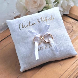 Puede incluir: Un cojín de anillos cuadrado de lino blanco con texto dorado "Christina & Roberto" y la fecha "6.07.2026". Dos anillos de boda dorados están atados con cintas blancas. El cojín está sobre una superficie de madera.