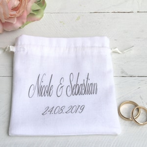 Könnte beinhalten: Ein weißer Baumwollbeutel mit Kordelzug, auf dem der Text "Nicole & Sebastian 24.08.2019" gedruckt ist. Zwei goldene Eheringe liegen auf einer weißen Holzoberfläche neben dem Beutel.