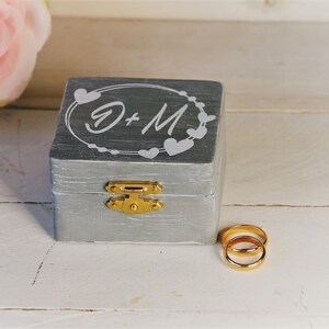 Könnte beinhalten: Graue Holz-Ringbox mit goldenem Verschluss und den Initialen "D + M" in einem herzförmigen Design. Zwei goldene Eheringe liegen neben der Box. Eine rosa Rose befindet sich im Hintergrund.