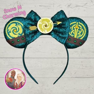 May include: Green and gold sequined Minnie Mouse ears with a bow and a gold charm featuring a swirling design and an arrow. The ears have a brown band with gold accents and the text "Snow N Charming".