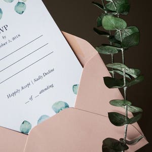 Op de afbeelding: Een roze envelop met een witte RSVP-kaart erin. De kaart heeft een aquarelontwerp met eucalyptusbladeren en de tekst "RSVP v&oacute;&oacute;r 5 oktober 2018".