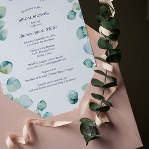 Op de afbeelding: Een lichtblauwe bruidsdoucheinvitatie met aquareleucalyptusbladeren en een roze envelop. De tekst van de uitnodiging luidt: "Kom gezellig naar een bruidsdouche ter ere van Audrey Annett Miller. Zaterdag 30 september 2018, 14:00 uur, Villages at the Domain, 11011 Domain Drive, Austin, Texas. RSVP v&oacute;&oacute;r 13 augustus aan Rachel (555) 333-5333, rachelgmail.com. Geregistreerd bij Bed Bath & Beyond | Pottery Barn | Target. Vul de bijgevoegde kaart in met je favoriete recept en breng het mee naar de bruidsdouche."