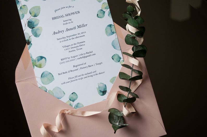 Op de afbeelding: Een lichtblauwe bruidsdoucheinvitatie met aquareleucalyptusbladeren en een roze envelop. De tekst van de uitnodiging luidt: "Kom gezellig naar een bruidsdouche ter ere van Audrey Annett Miller. Zaterdag 30 september 2018, 14:00 uur, Villages at the Domain, 11011 Domain Drive, Austin, Texas. RSVP v&oacute;&oacute;r 13 augustus aan Rachel (555) 333-5333, rachelgmail.com. Geregistreerd bij Bed Bath & Beyond | Pottery Barn | Target. Vul de bijgevoegde kaart in met je favoriete recept en breng het mee naar de bruidsdouche."