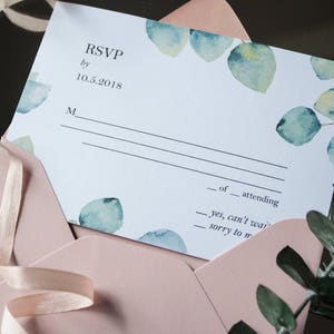 Op de afbeelding: Een witte RSVP-kaart met een aquarelontwerp van groene bladeren. De kaart heeft de tekst "RSVP by 10.5.2018" en een regel voor de naam van de gast.