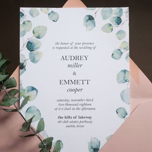 Op de afbeelding: Huwelijksuitnodiging met een witte achtergrond en een aquarelontwerp van eucalyptusbladeren in tinten groen en blauw. De tekst luidt "the honor of your presence is requested at the wedding of Audrey miller & Emmett cooper saturday, november third two thousand eighteen at 2 o'clock in the afternoon the hills of lakeway 26 club estates parkway austin, texas"