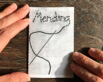May include: A white fabric rectangle with the word "Mending" embroidered in black thread. A black thread and needle are stitched onto the fabric, suggesting a sewing or repair theme. The image is held by two hands.