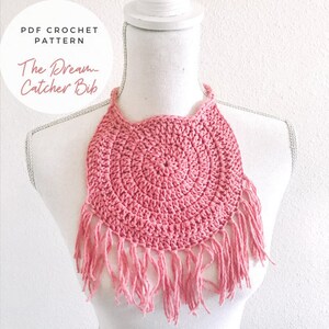 May include: A pink crocheted bib with a fringe border. The bib is shaped like a circle with a smaller circle in the center. The text "The Dream-Catcher Bib" is visible in the image.