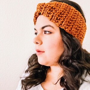 May include: A brown crocheted headband with a twisted knot design. The headband is worn on a person with long, dark brown hair.