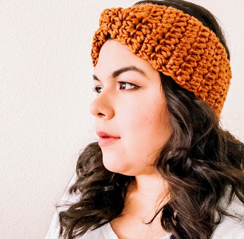 May include: A brown crocheted headband with a twisted knot design. The headband is worn on a person with long, dark brown hair.