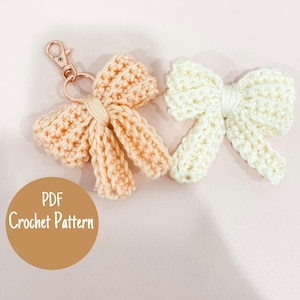 May include: Two crocheted bow keychains, one peach and one cream, are shown on a light pink surface. The peach bow has a gold-coloured clasp. The text "PDF Crochet Pattern" is in a brown circle.