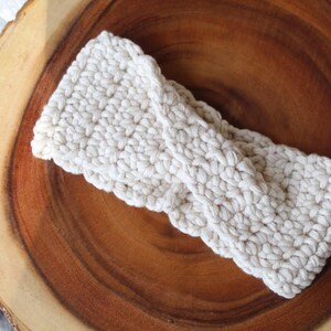 May include: A white crocheted headband with a twisted knot design. The headband is on a wooden surface.