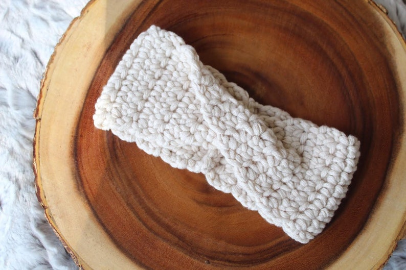 May include: A white crocheted headband with a twisted knot design. The headband is on a wooden surface.