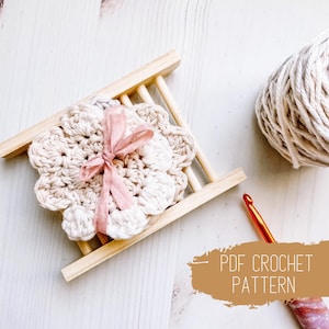 May include: A crochet flower coaster in a natural colour with a pink ribbon tied around it. The coaster is sitting on a wooden soap dish. There is a ball of yarn and a crochet hook in the background. The text "PDF CROCHET PATTERN" is written on a brown background.
