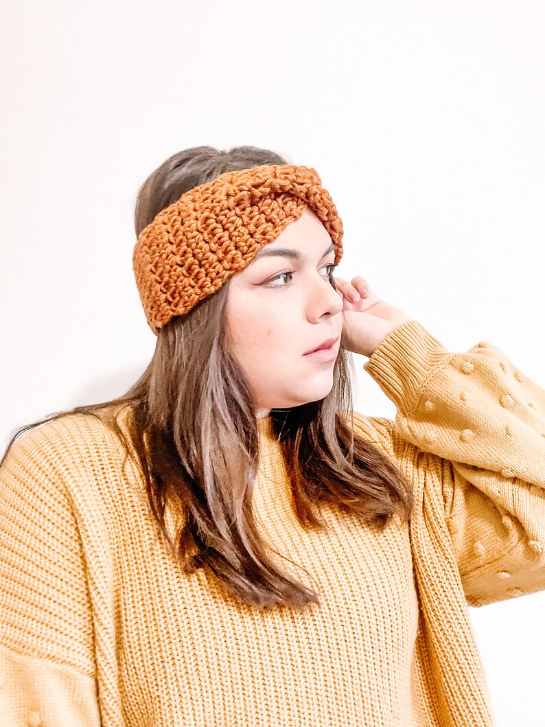 May include: A brown crocheted headband with a twisted design. The headband is worn by a person with long brown hair and a yellow sweater.