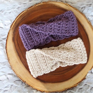 May include: Two crocheted headbands, one purple and one cream, displayed on a wooden slice.