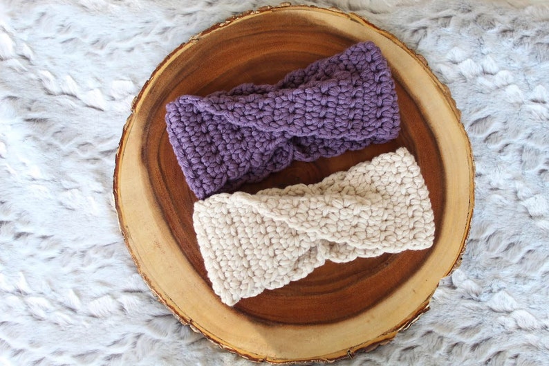 May include: Two crocheted headbands, one purple and one cream, displayed on a wooden slice.