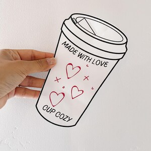 May include: White cup cozy with black outline, featuring red hearts and the text "MADE WITH LOVE" and "CUP COZY". The design resembles a coffee cup, ideal for keeping beverages warm.