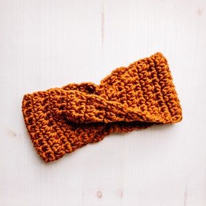 May include: A brown crocheted headband with a twisted design. The headband is made of a soft, textured yarn.
