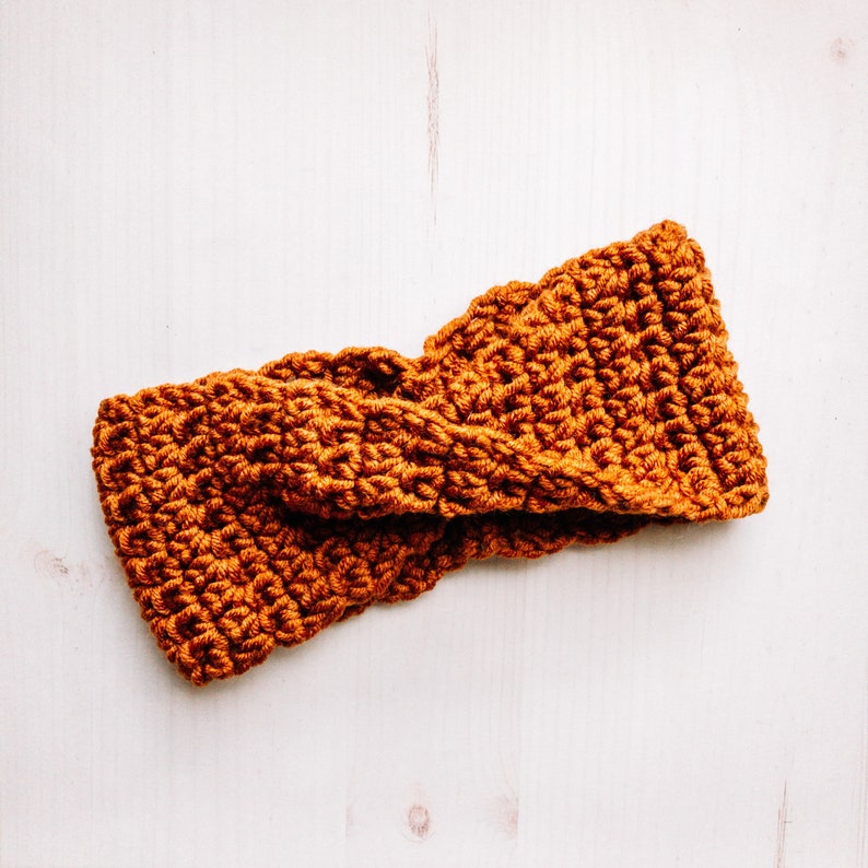 May include: A brown crocheted headband with a twisted design. The headband is made of a soft, textured yarn.