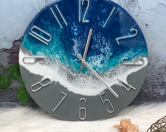 May include: A round wall clock featuring a resin art design that mimics an ocean shoreline. The top half displays deep blue and turquoise water with white foam waves, while the bottom half is solid grey. It has silver-toned metal numerals.