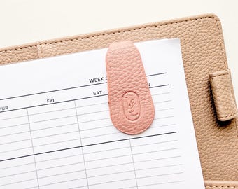 Può includere: Un fermaglio segnapagina in pelle rosa chiaro con un design impresso, attaccato a una pagina dell'agenda. La clip è sopra un foglio bianco con un programma settimanale. L'agenda è di colore beige chiaro.