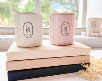 Può includere: Due candele cilindriche, una color crema e una rosa, sono appoggiate su una pila di tre libri. Ogni candela ha un logo nero. I libri sono in tonalità di rosa e nero. Lo sfondo mostra una finestra con una vista sfocata di alberi.