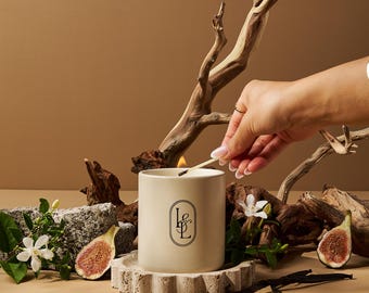 Può includere: Una candela beige accesa in un portacandele in ceramica con un logo nero. La candela è circondata da fichi, baccelli di vaniglia e legno alla deriva. Una mano accende la candela con un fiammifero. La candela poggia su una base di pietra.