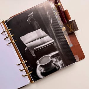 May include: Brown leather planner with gold-colored rings and a pen. The planner has a black and white image inside. The planner has a brown leather strap with a logo. The planner has monthly tabs.
