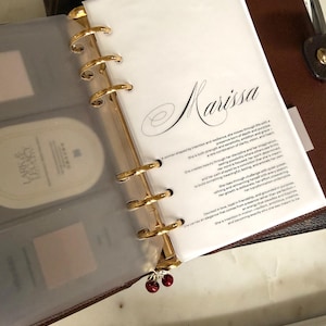 May include: A brown leather planner with gold-colored rings and a cherry charm. The planner is open to a white page with the name "Marissa" in elegant script and additional text. A clear plastic sleeve is visible.
