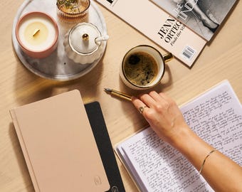 Può includere: Una composizione piatta con una candela accesa in un contenitore rosa, un vassoio di marmo con fiori, una tazza di caffè, una rivista e due quaderni. Una mano con una penna d'oro scrive in un quaderno aperto.