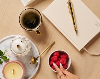 Può includere: Vista dall'alto di una composizione serena con una tazza di caffè, un taccuino, una penna d'oro, una piccola ciotola di lamponi, una candela e una zuccheriera su un vassoio di marmo. Gli oggetti sono disposti su una superficie di legno chiaro.