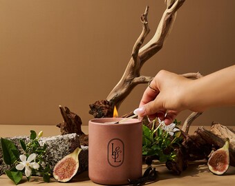 Può includere: Una candela rosa accesa con un logo nero è circondata da fichi, fiori bianchi e legno. Una mano accende la candela con un fiammifero. La candela è in un contenitore cilindrico.