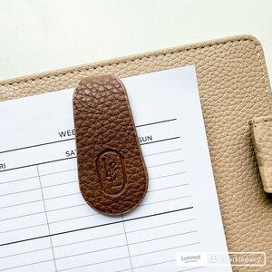 May include: A brown leather bookmark with a debossed logo, resting on a white planner page. The bookmark has a textured surface and is positioned over the days of the week. The planner is a light beige color.