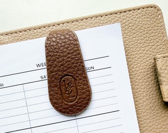 Può includere: Un segnalibro in pelle marrone con logo impresso, appoggiato su una pagina di agenda bianca. Il segnalibro ha una superficie strutturata ed è posizionato sui giorni della settimana. L'agenda è di colore beige chiaro.