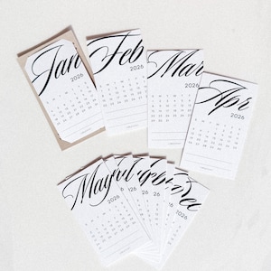 May include: A collection of small, white 2026 calendars, each featuring a different month. The month names are written in elegant black script. The calendars are arranged on a white surface, with the month of January on a tan card.